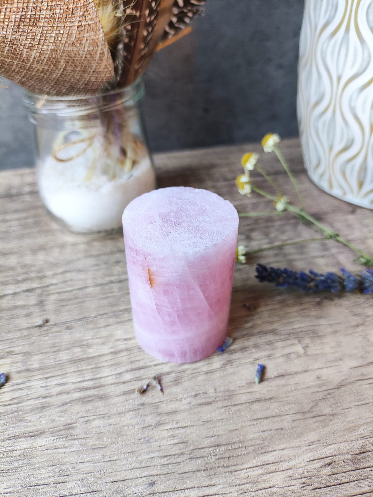 Cilindro de cuarzo rosa pulido sobre mesa de madera, acompañado de flores secas y lavanda, ambientación rústica y esotérica.
