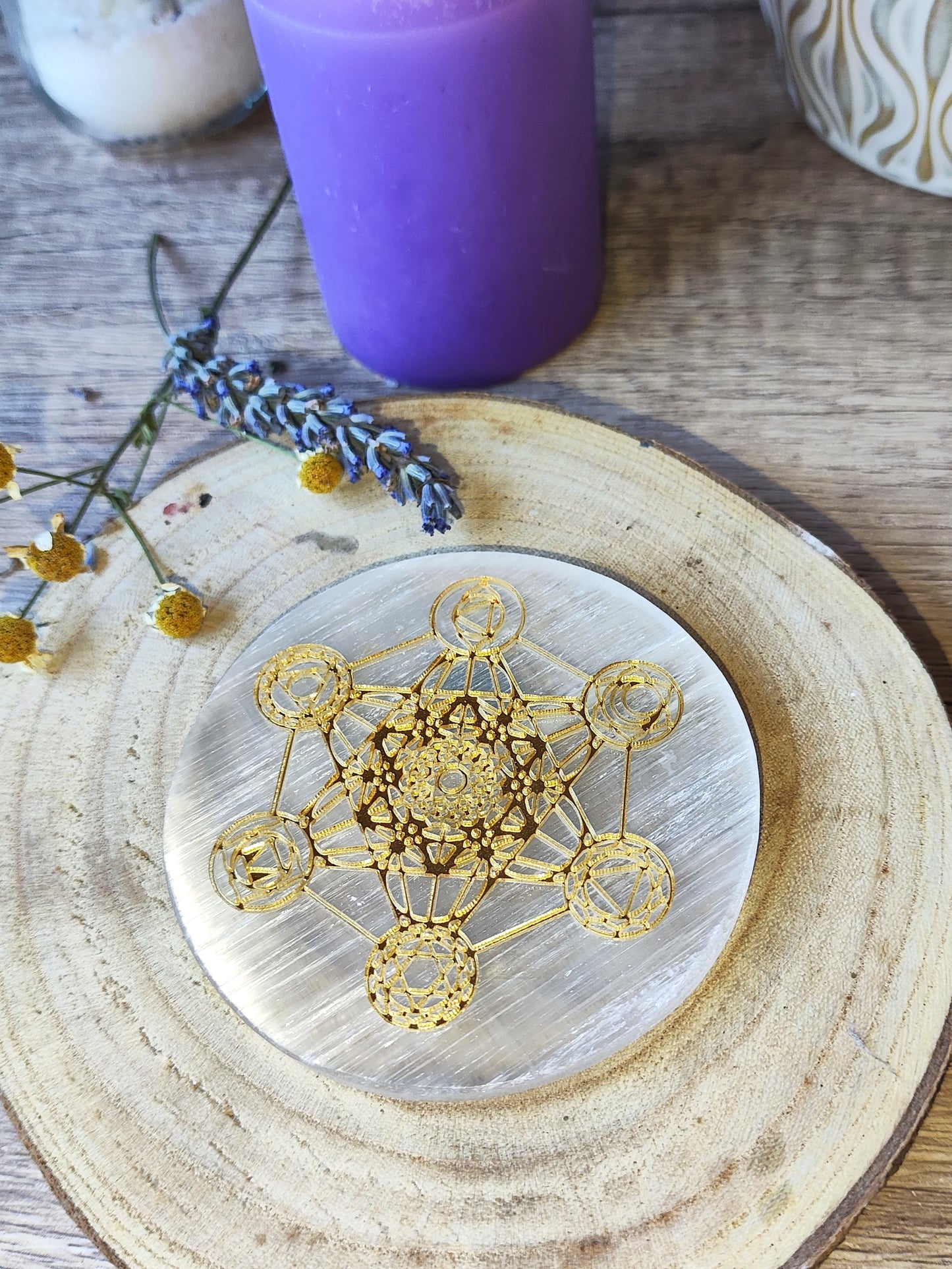 Placa redonda de selenita con Cubo de Metatrón dorado sobre base de madera, con vela y lavanda como ambientación esotérica.

