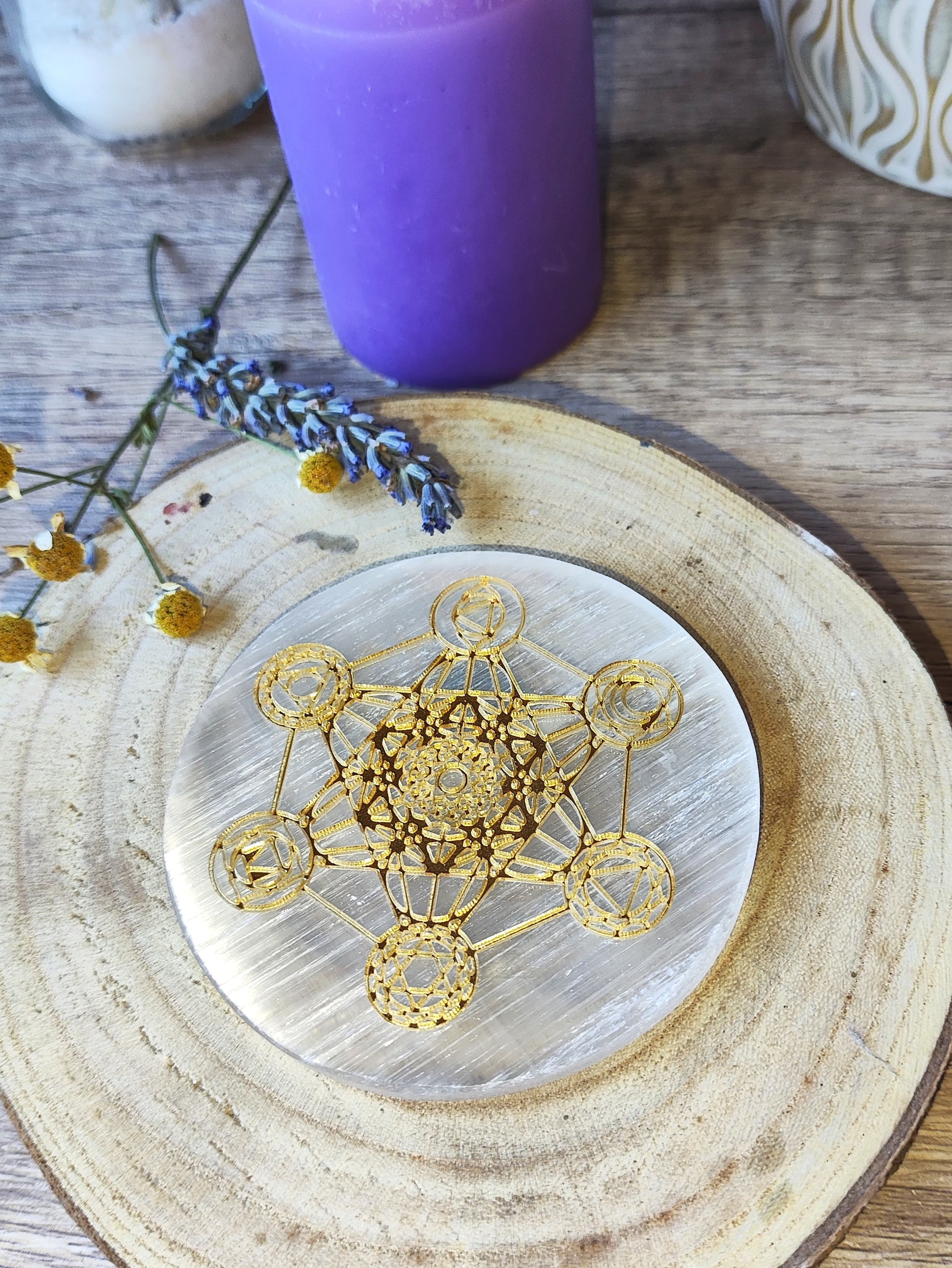 Placa redonda de selenita con Cubo de Metatrón dorado sobre base de madera, con vela y lavanda como ambientación esotérica.

