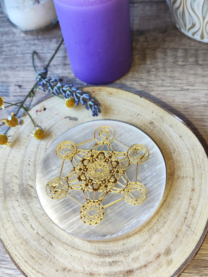 Placa redonda de selenita con Cubo de Metatrón dorado sobre base de madera, con vela y lavanda como ambientación esotérica.

