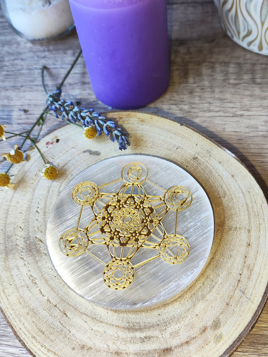 Placa redonda de selenita con Cubo de Metatrón dorado sobre base de madera, con vela y lavanda como ambientación esotérica.

