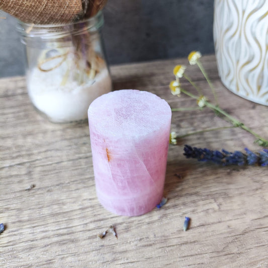 Cilindro de cuarzo rosa pulido sobre mesa de madera, acompañado de flores secas y lavanda, ambientación rústica y esotérica.

